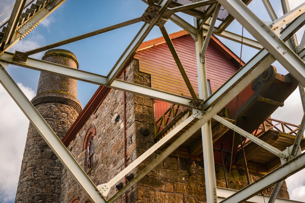 A close-up view of a historic stone building with a cylindrical chimney, seen through the geometric metal framework of a bridge or industrial structure against a blue sky.