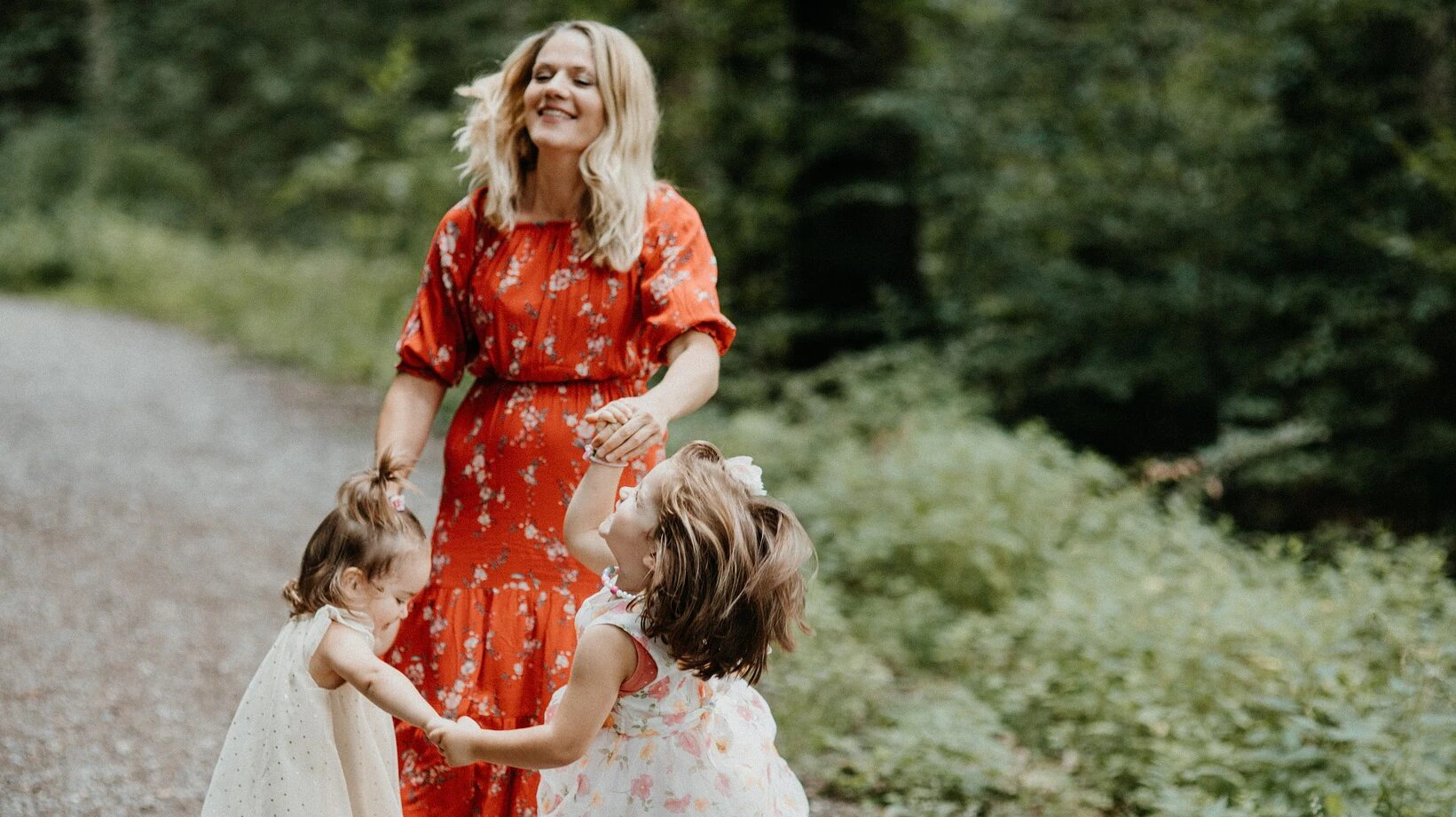 A woman in a red dress smiles as she holds hands and dances in a circle with two young girls in light-colored dresses on a gravel path surrounded by greenery.