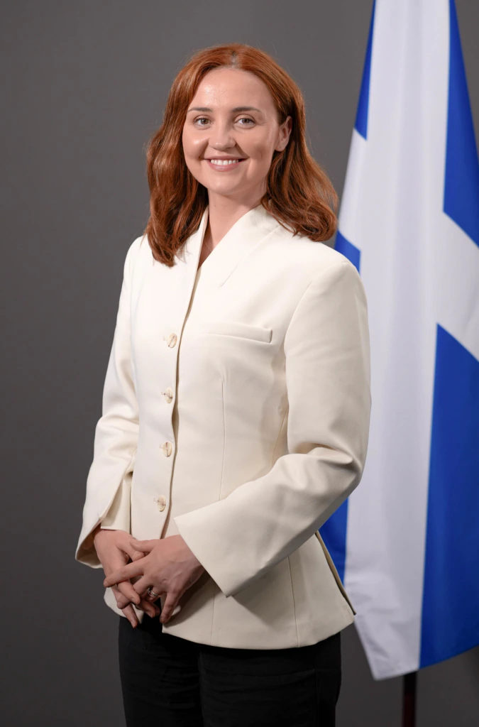 A woman with shoulder-length red hair wearing a white blazer stands in front of a grey background next to a flag with blue and white colors. She is smiling and has her hands loosely clasped.