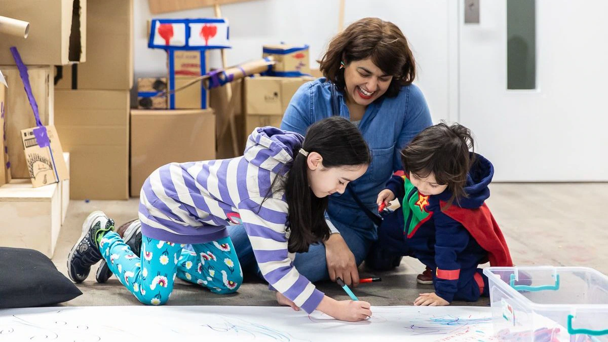 A woman smiles while watching two young children draw on a large sheet of paper on the floor. The children are engaged in drawing, and the room has cardboard structures and art supplies in the background.