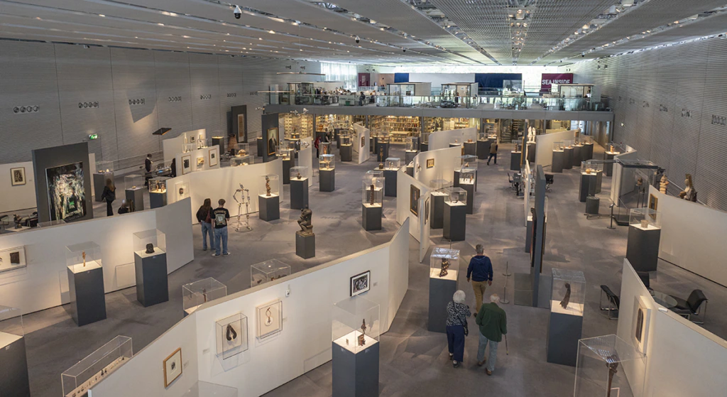 A spacious, modern art museum with high ceilings displays sculptures and framed artworks in glass cases and on white walls. Visitors walk and observe the exhibits along wide, curved paths.