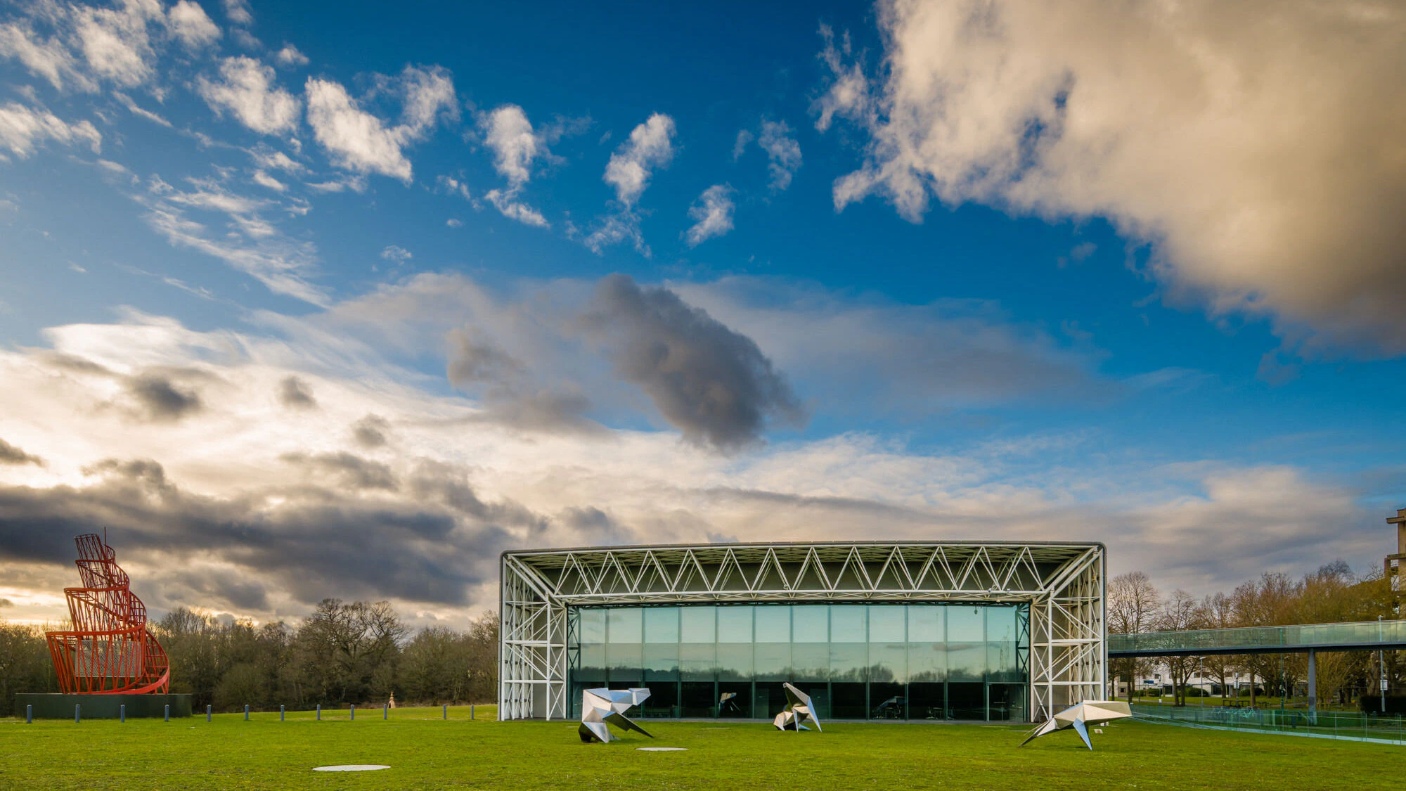 A modern glass building with a white steel frame stands on a green lawn under a partly cloudy sky. Sculptures are scattered on the grass, and a red spiral structure is visible on the left side of the scene.