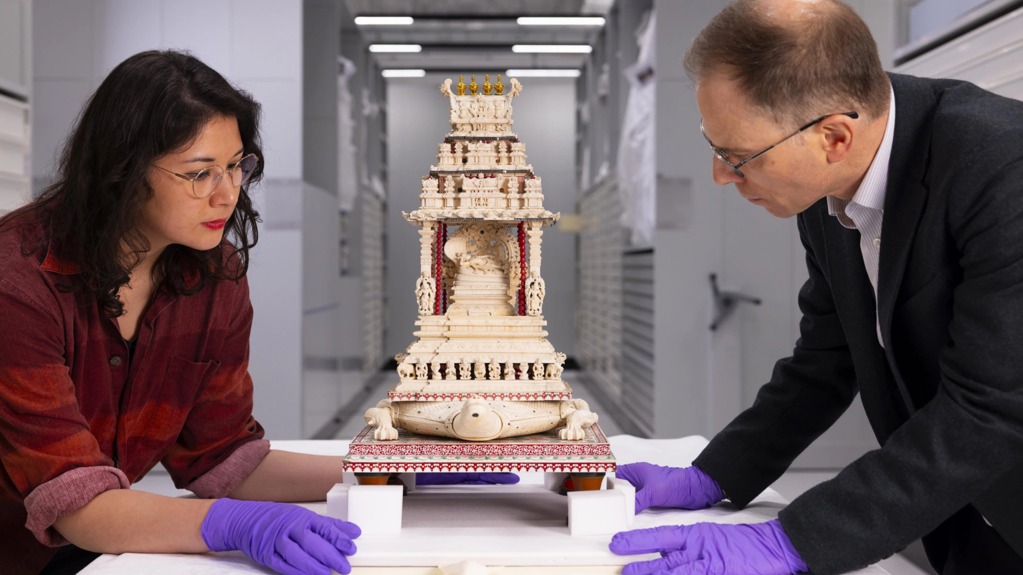 Two people wearing purple gloves carefully examine an ornate, ivory-colored miniature temple with intricate carvings, inside a modern, well-lit storage or conservation facility.