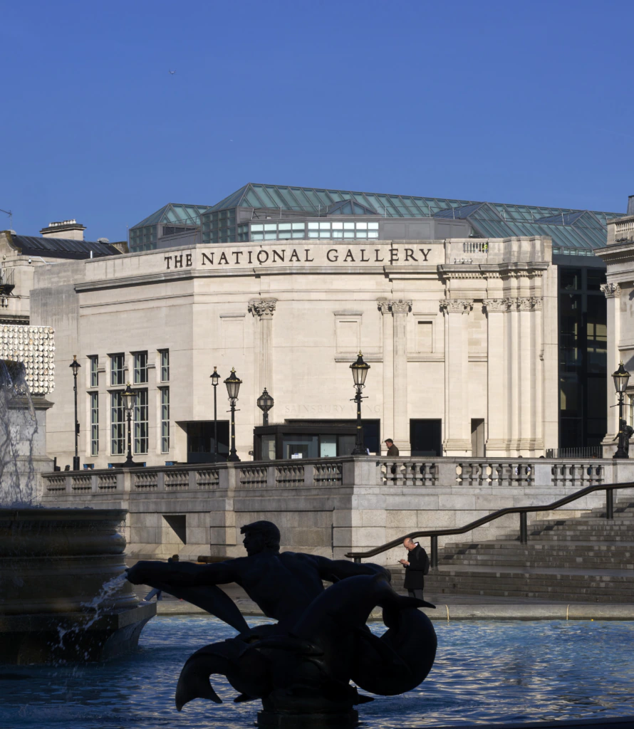 The National Gallery building in London, with its grand white facade, seen behind a fountain and a silhouetted statue under a clear blue sky.