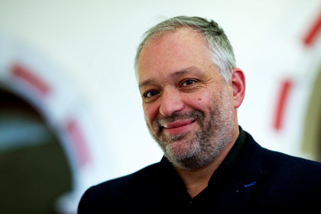 A middle-aged man with short gray hair and a beard, wearing a dark jacket, smiles at the camera against a blurred background with white and red shapes.