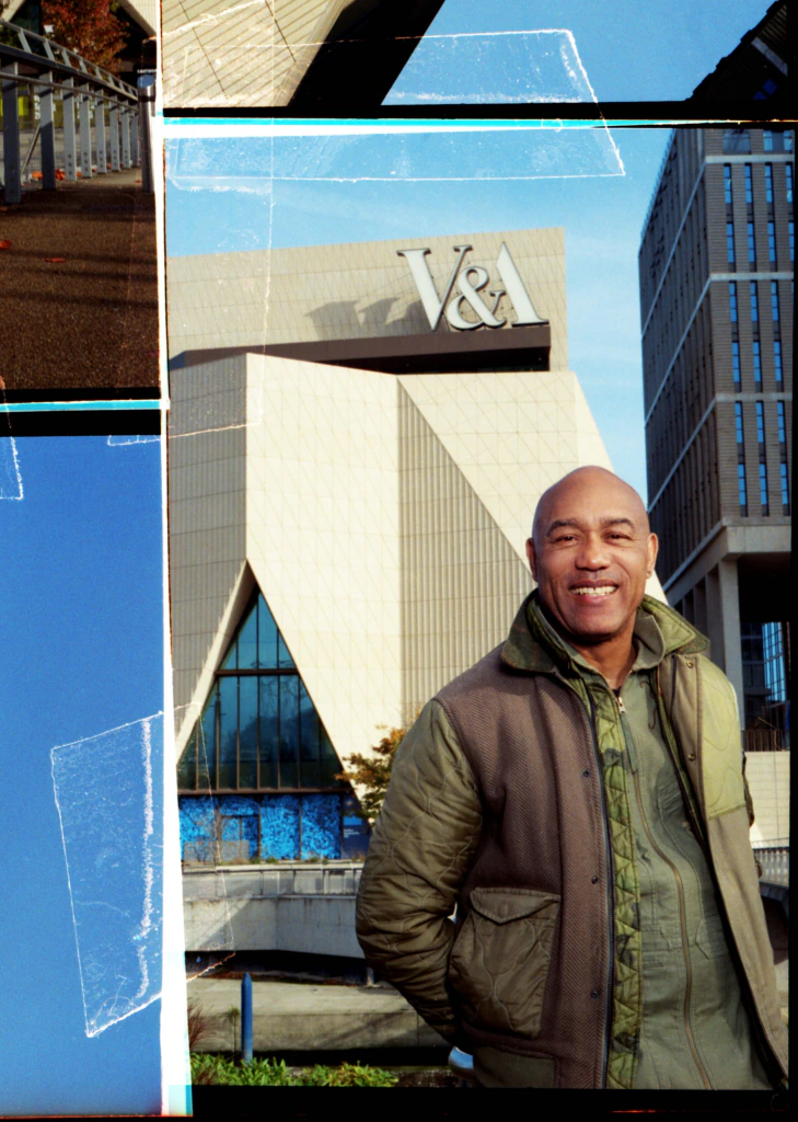 A man wearing a green jacket smiles while standing in front of the V&A Museum. The building has a modern, geometric facade and large V&A letters on top. The image has film borders and light leaks.