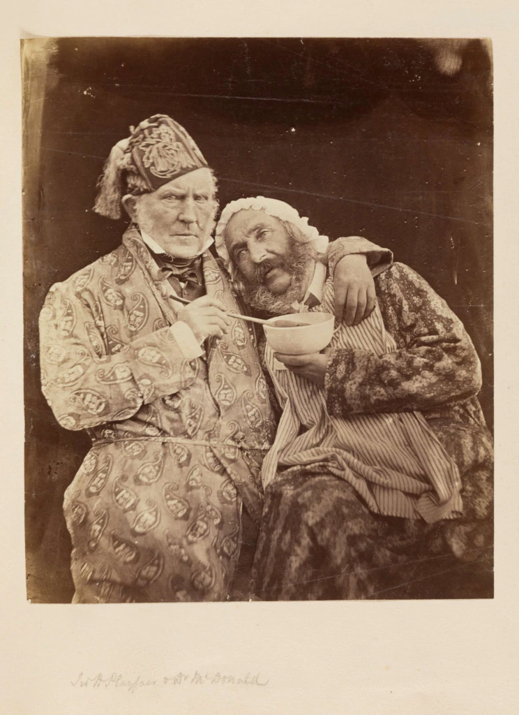 Two men dressed in ornate, patterned robes pose closely together. One wears a patterned hat and looks stern, while the other, with a headscarf and beard, holds a bowl and smiles, resting his arm on the first mans shoulder.