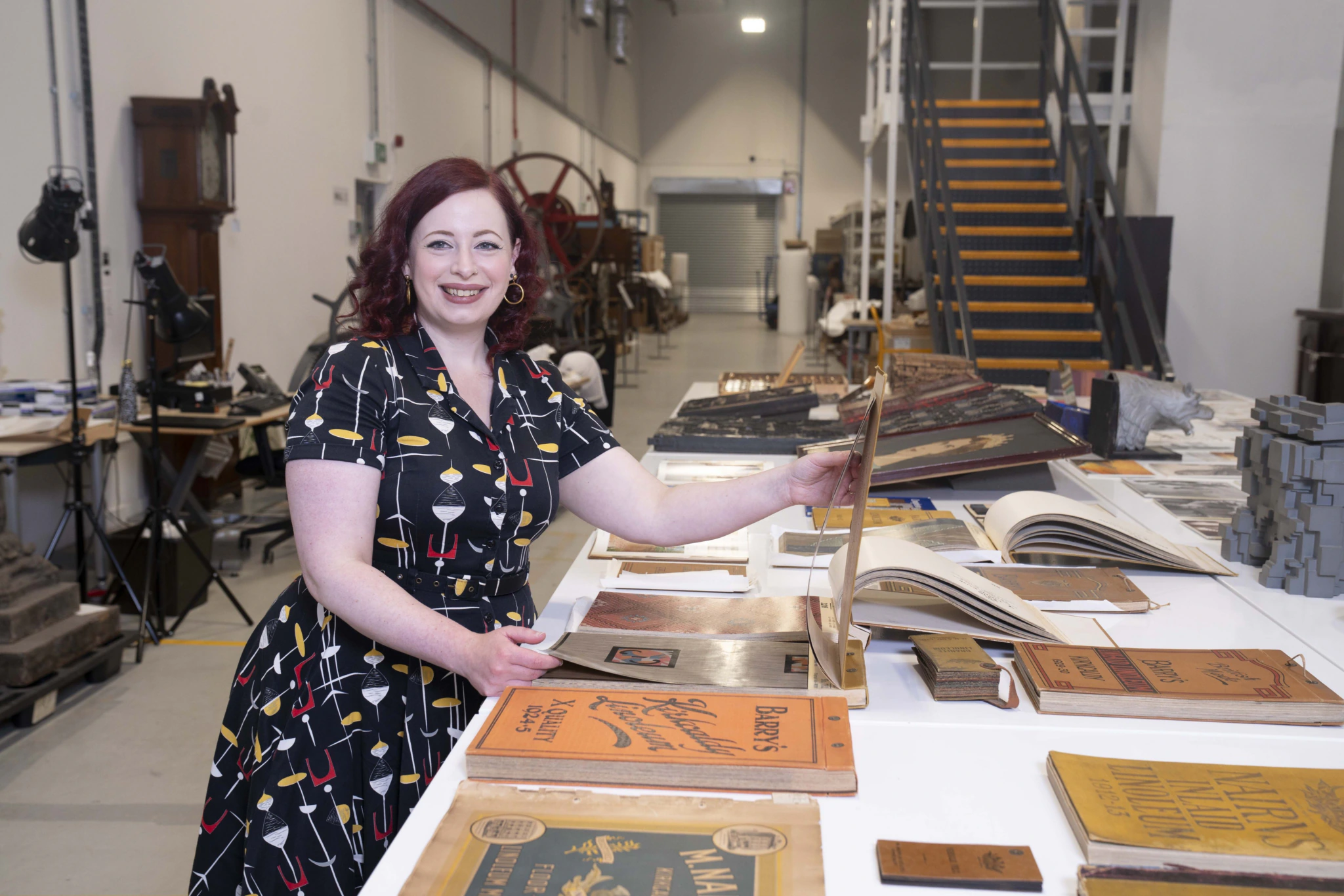 A smiling woman with red hair and a patterned dress stands at a large table covered with vintage books and documents in a spacious, industrial-style room with shelves and equipment in the background.