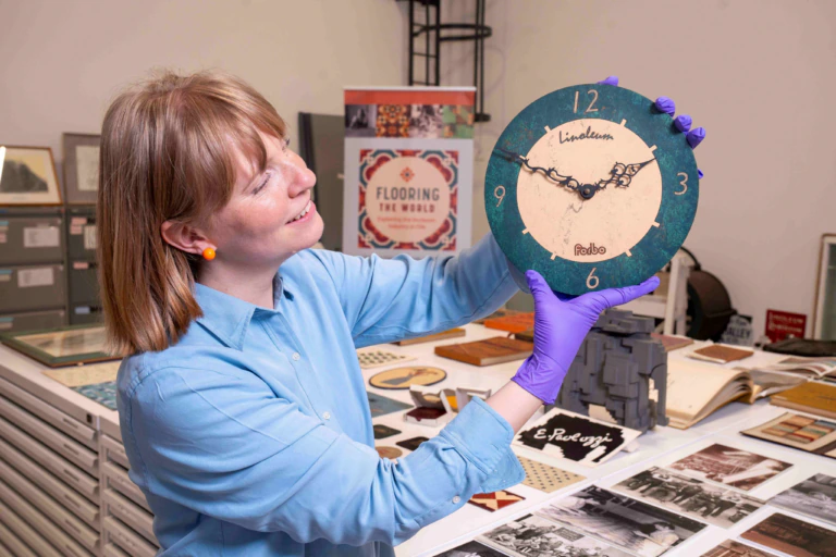 A woman wearing a blue shirt and purple gloves holds up a vintage wall clock and smiles. She stands in a room filled with historical artifacts, documents, and photos related to flooring.
