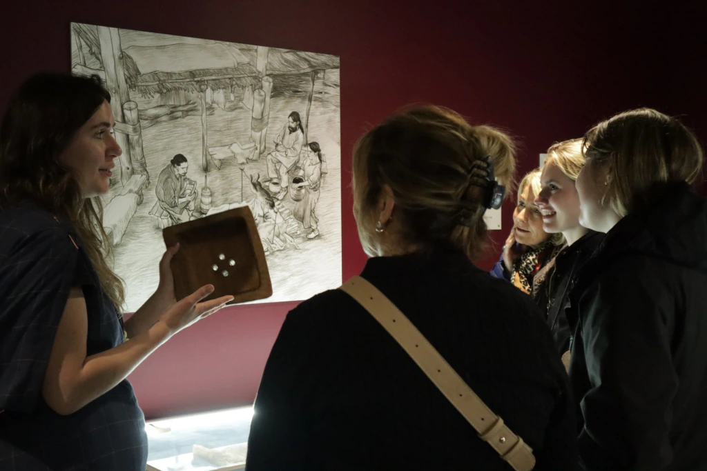 A woman holds up an artifact and speaks to a small group of people in a museum. Behind them is a black-and-white drawing of people working. The group listens attentively in the dimly lit space.