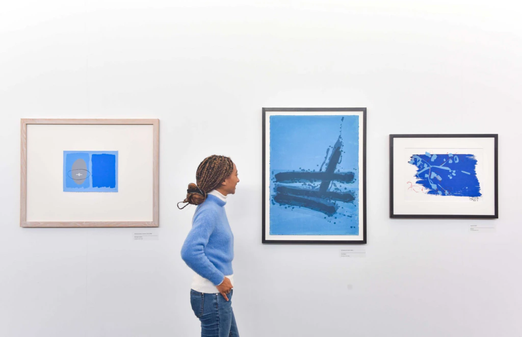 A woman in a blue sweater and jeans views three framed abstract blue artworks displayed on a white gallery wall.