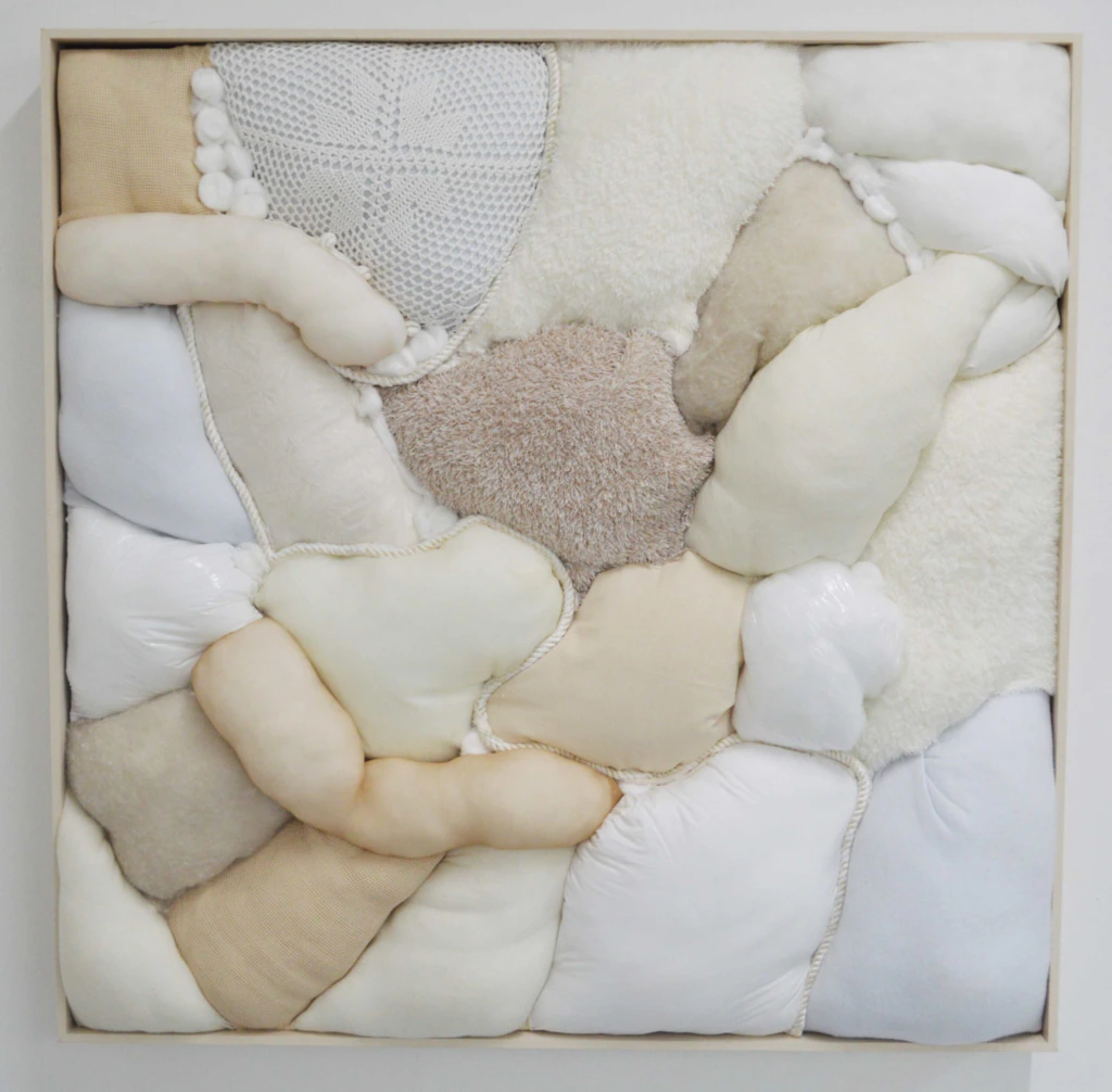 A soft, padded textile artwork features irregular, rounded shapes in various neutral tones—white, beige, taupe, and cream—stitched together within a square frame, creating a plush, quilted appearance with varied textures.