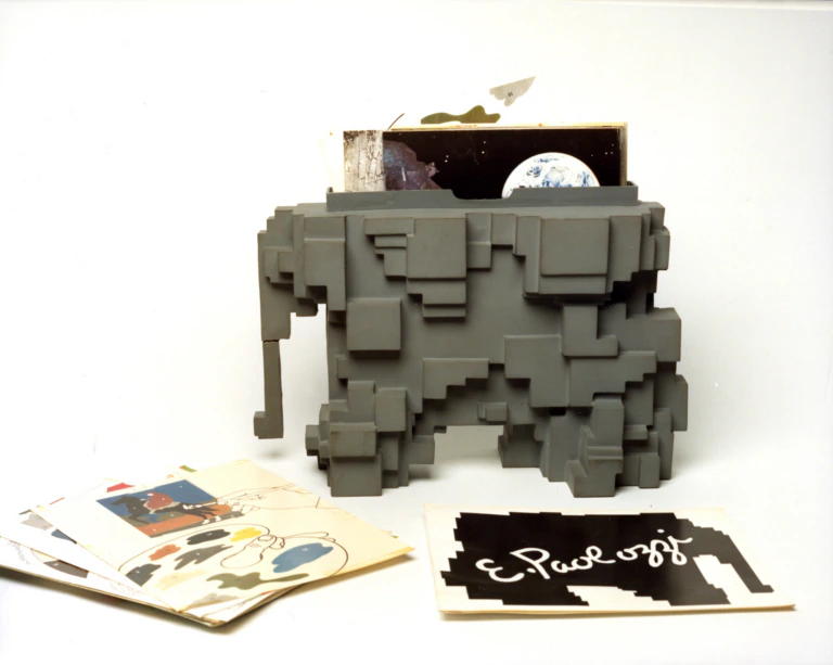 A geometric, gray, sculpture-like file holder is filled with papers, including an image of Earth from space. Assorted colorful papers and a card with E. Paoluzzi written on it are spread out in front of the holder.