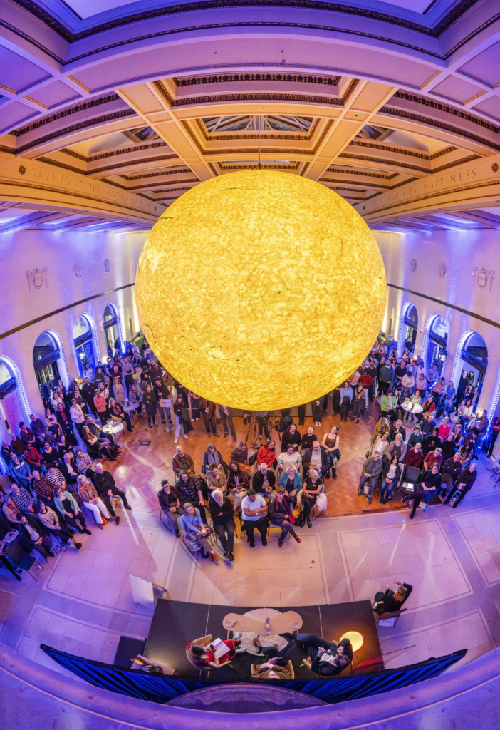 A large, glowing, yellow sphere resembling the sun hangs above a crowd of people gathered in a grand hall with ornate ceilings and arched windows, creating a vibrant, immersive atmosphere.