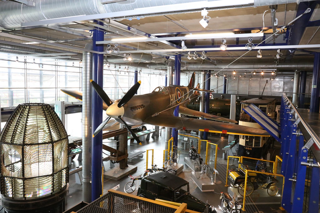 A vintage fighter plane is displayed suspended in a modern museum with glass walls, blue beams, bicycles, and various historical vehicles. Industrial lighting and a large lens exhibit are also visible.