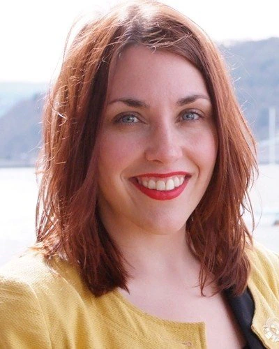 A woman with shoulder-length brown hair, blue eyes, and red lipstick smiles at the camera. She is wearing a yellow jacket and is outdoors, with a blurred background of water and hills.