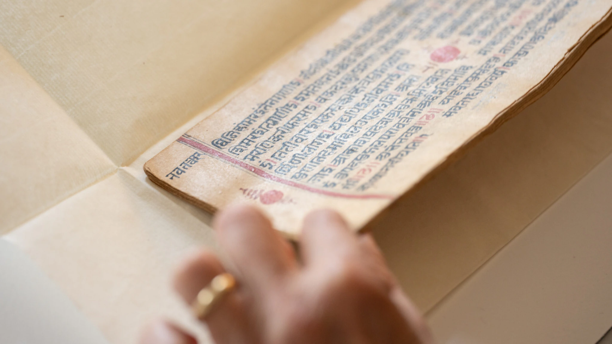 A hand gently turns the page of an old manuscript with text in Sanskrit script, written in black and red ink on aged paper.