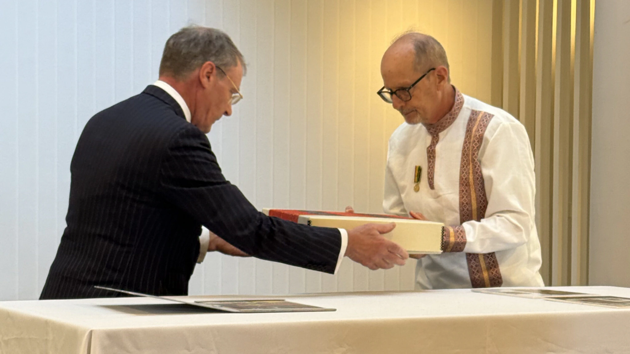 Two men stand across a table; one in a striped suit hands a boxed document to the other, who wears glasses and a traditional embroidered white shirt. Both appear focused and solemn during the exchange.