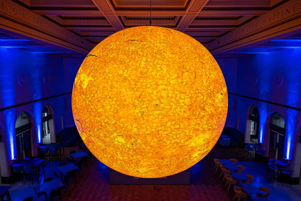 A large, illuminated model of the Sun glows bright yellow-orange in the center of a dimly lit room with blue walls, high ceilings, arched windows, and empty tables and chairs arranged below.
