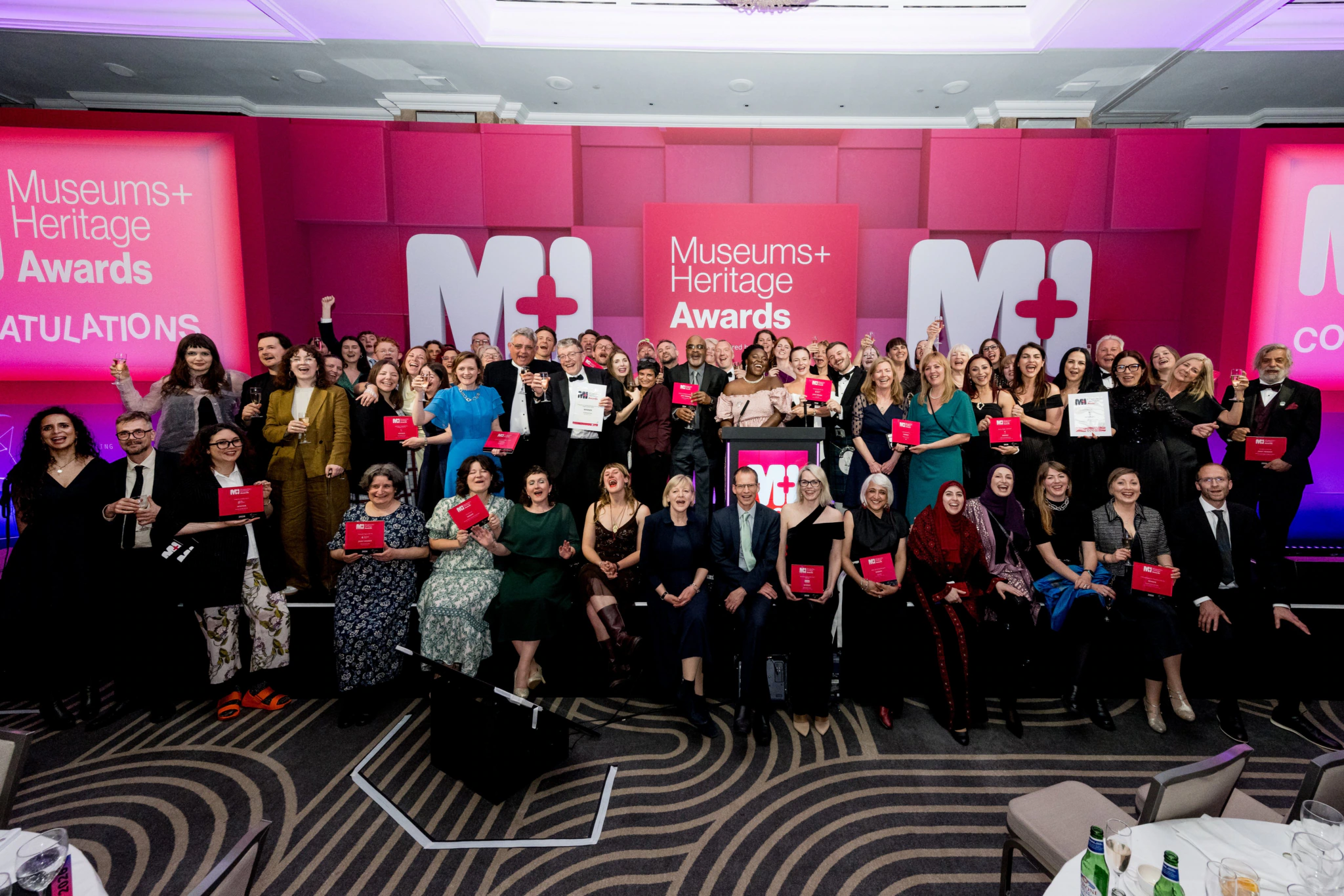 A large group of people pose together on stage holding awards at the Museums + Heritage Awards, with pink and white event signage in the background and attendees dressed formally, smiling and celebrating.