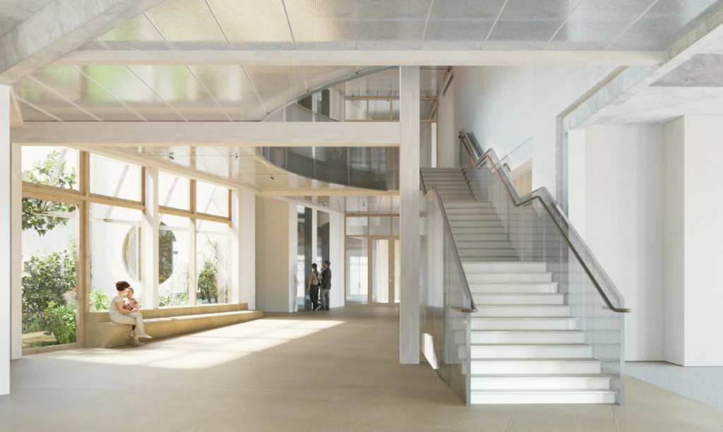 A bright, modern building interior with large windows, an open staircase, and a person sitting on a bench by the windows. Two people are seen talking in the background near a glass wall. The space is filled with natural light.