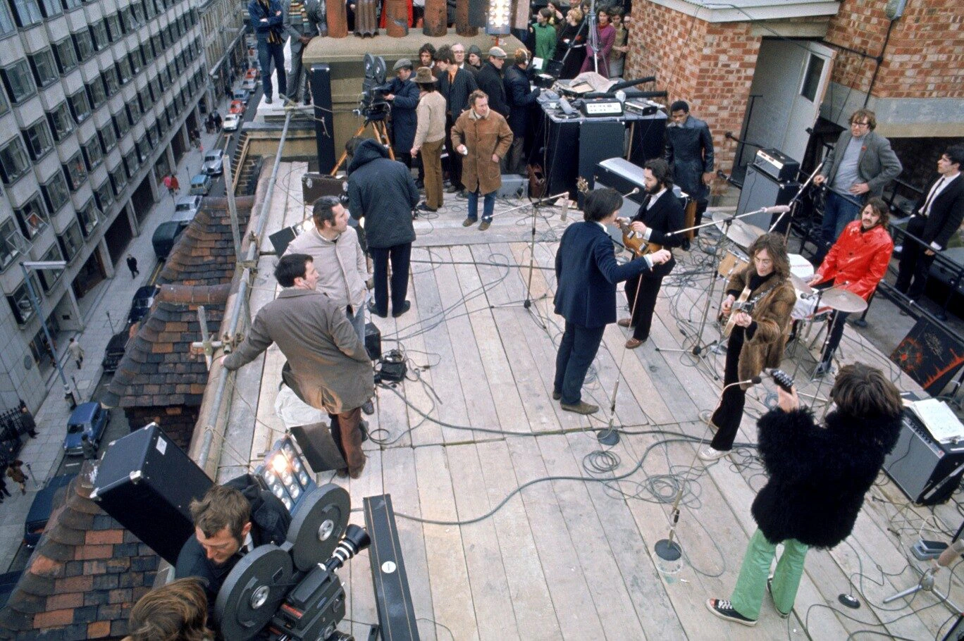 A band performs on a rooftop stage surrounded by musical equipment and cameras, with several people watching and city buildings visible in the background.