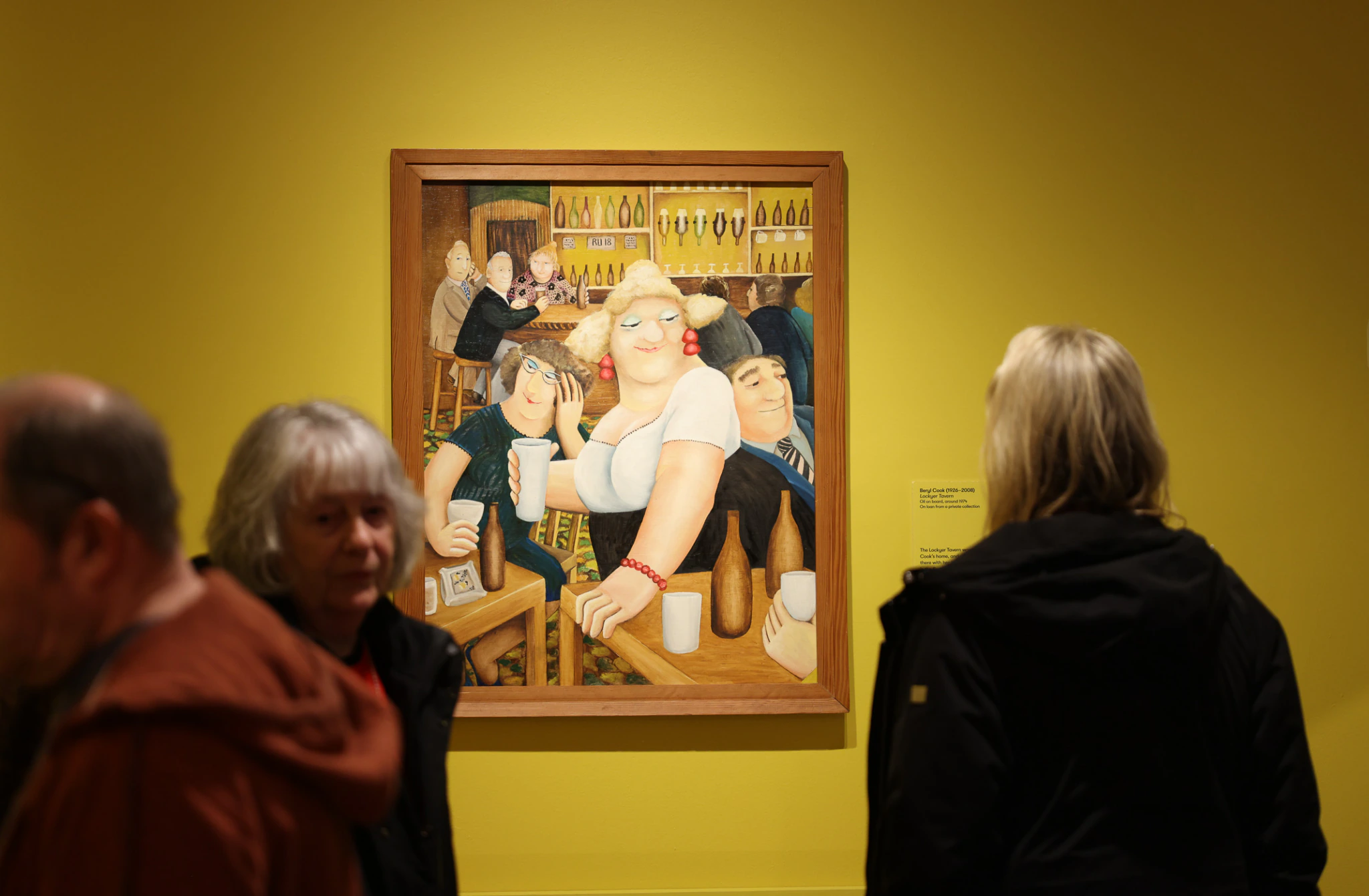 People observe a colorful painting of a lively bar scene displayed on a yellow wall in an art gallery. Several viewers stand in the foreground, their backs turned toward the artwork.