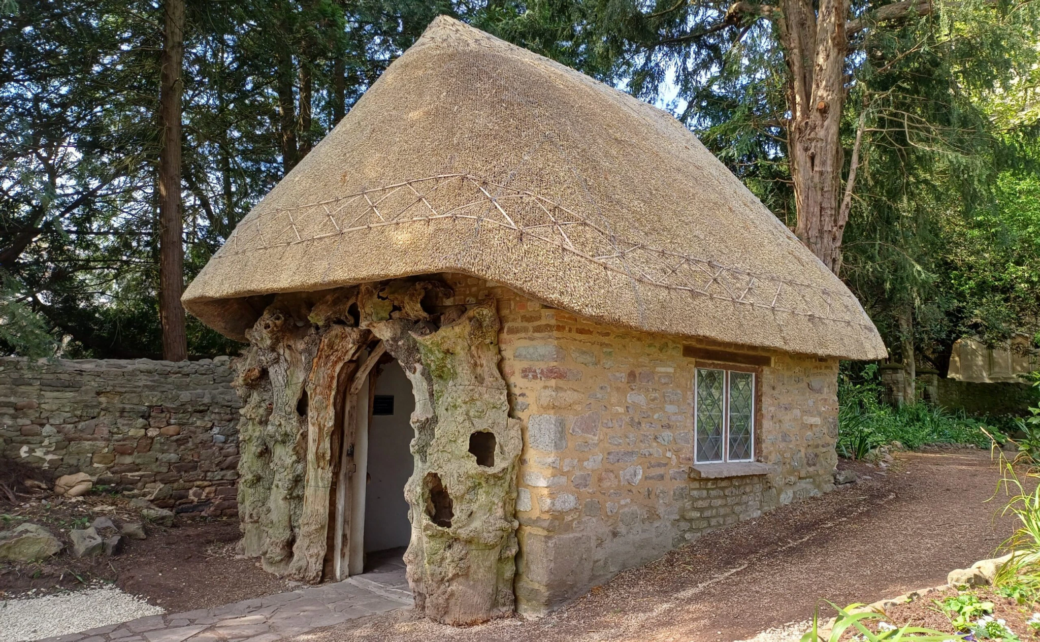 A small stone cottage with a thatched roof, rustic wooden doorframe, and one window, surrounded by trees and greenery on a sunny day.