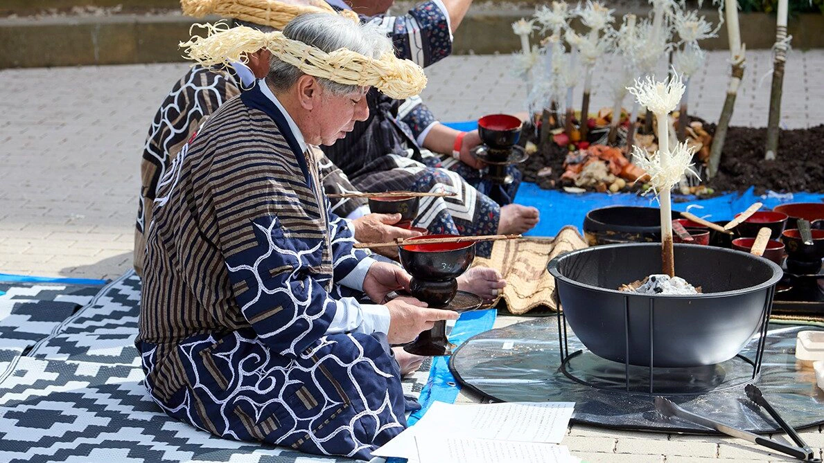Three people in traditional Japanese attire and straw headbands sit on patterned mats outdoors, performing a ritual with wooden bowls and food offerings arranged near white decorative branches.