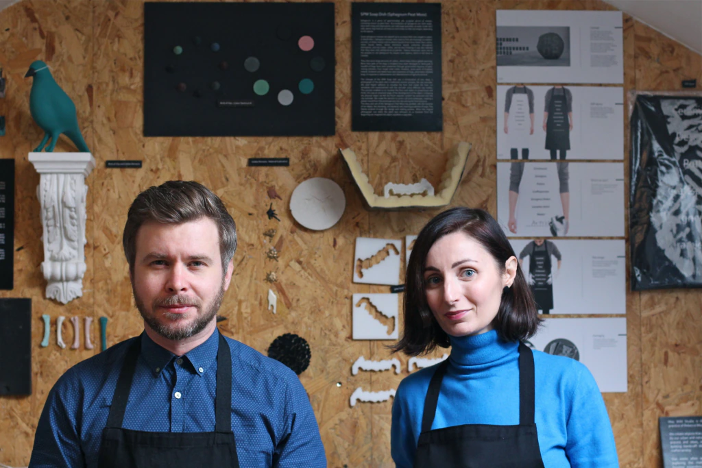 A man and a woman wearing black aprons stand side by side in front of a wooden wall decorated with various art pieces and informational posters. Both look toward the camera with neutral expressions.