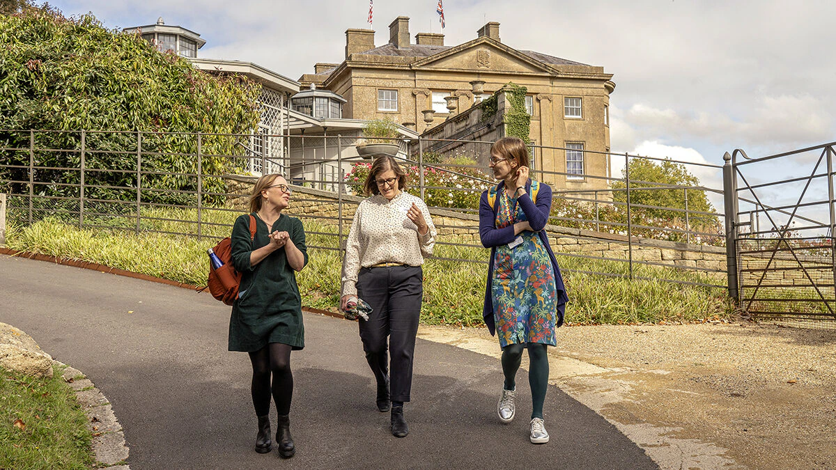 Three women walk and chat on a paved path in front of a large historic building with flags on the roof, surrounded by greenery on a partly cloudy day.