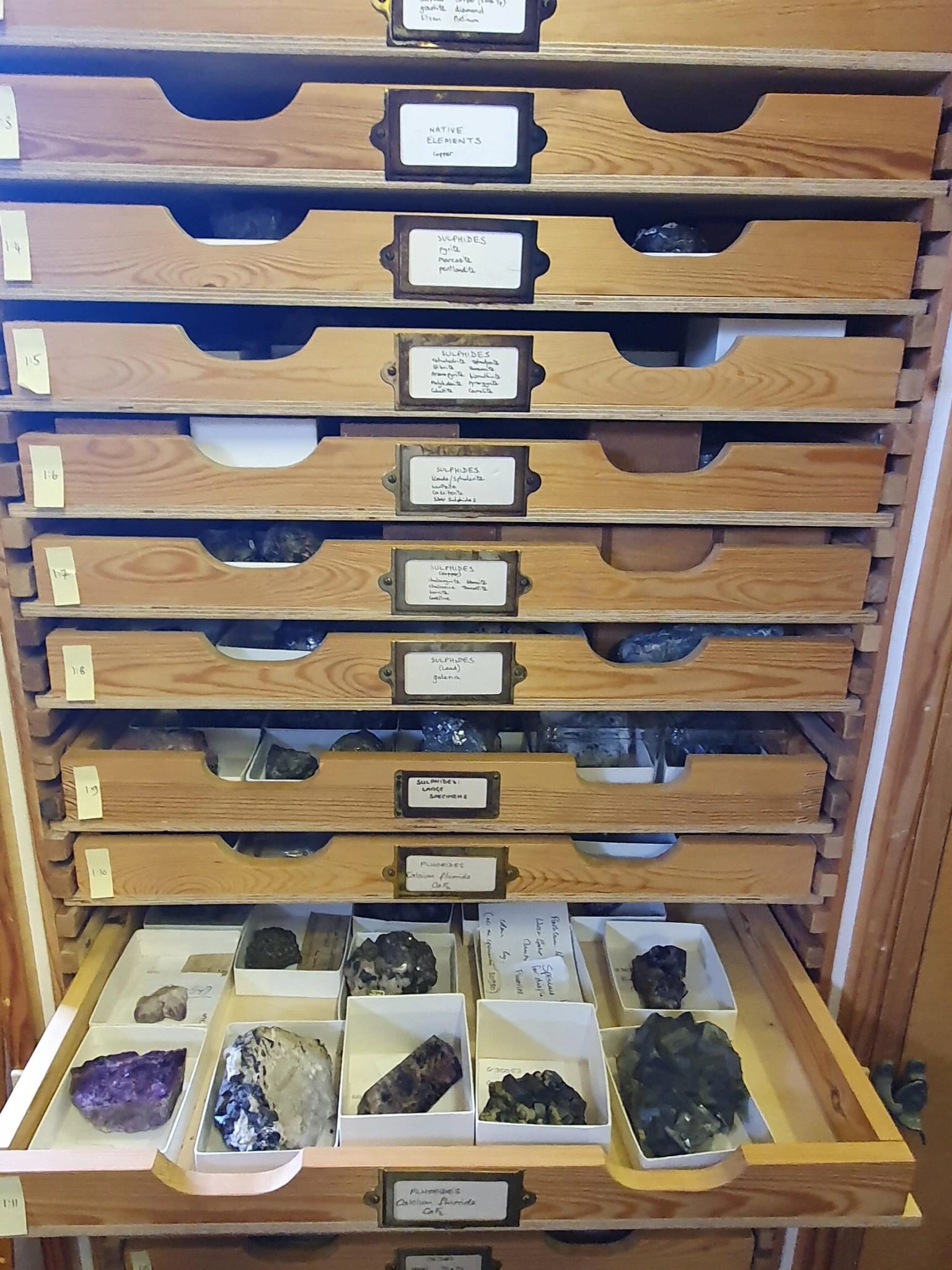 A wooden cabinet with several open drawers, each containing labeled compartments filled with various mineral and rock specimens, some visibly colorful and distinct in shape.