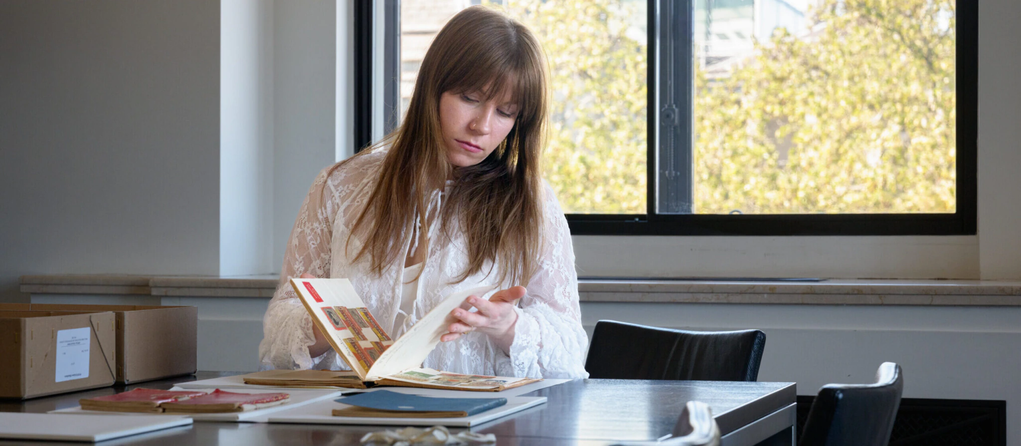 A woman with long brown hair sits at a table by a window, reading a book. She is surrounded by books and papers, with trees and buildings visible outside.