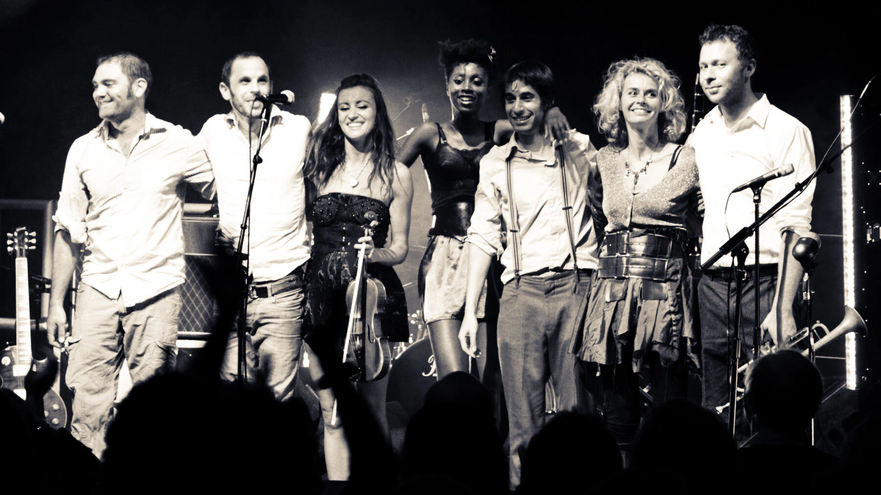 A group of seven musicians stand side by side on stage, smiling with arms around each other, in front of musical instruments and equipment, as an audience looks on. The image is in black and white.