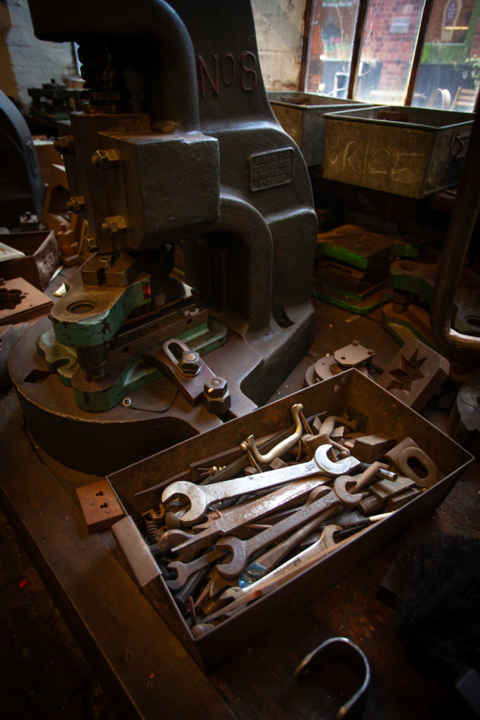 A box of assorted wrenches sits on a workbench next to a large industrial machine in a workshop with scattered tools and worn surfaces. Sunlight streams through dusty windows in the background.