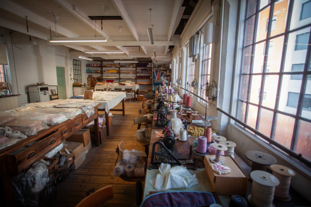 A spacious, sunlit textile workshop with sewing machines, spools of thread, fabric rolls, and worktables scattered throughout the wooden-floored room. Large windows line one wall, letting in plenty of natural light.