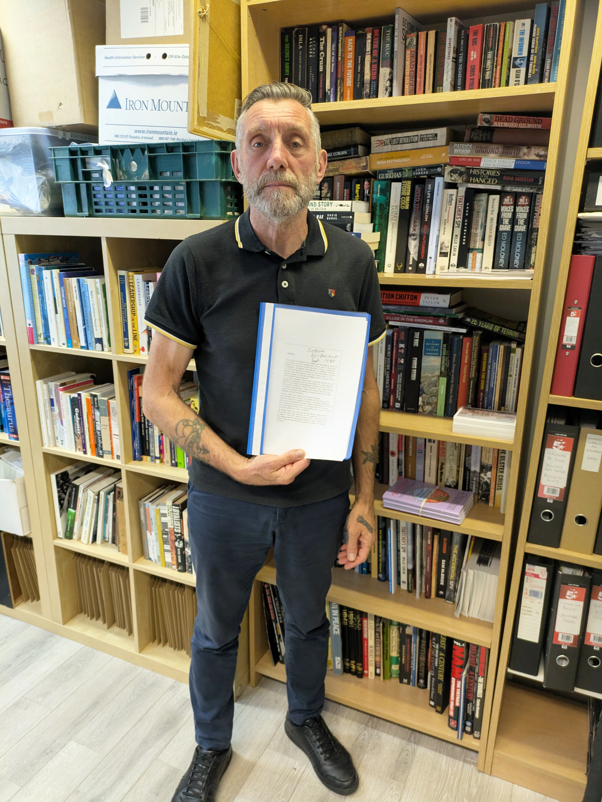 A man with tattoos, wearing a black polo shirt and blue pants, stands in front of bookshelves filled with books, holding up a document in a blue folder.