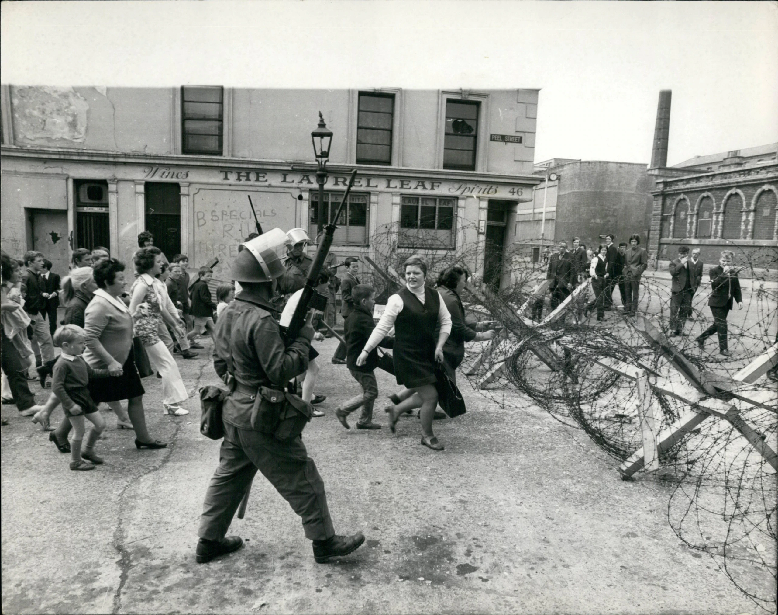 A soldier with a rifle stands by barbed wire as a crowd, including women and children, walks past on a city street near a building called The Laurel Leaf. Some people look at the soldier; the mood appears tense.