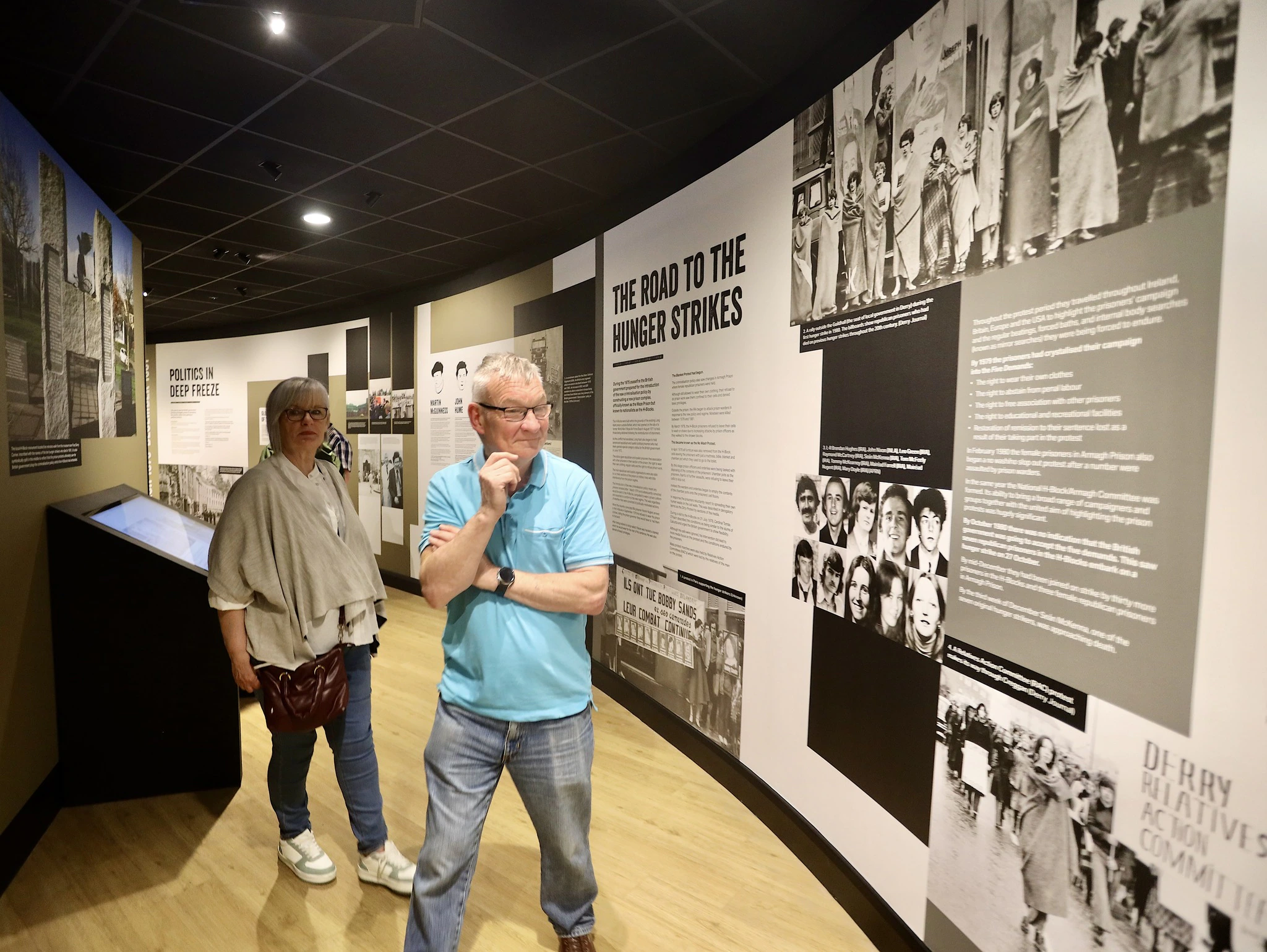 Two people walk through a museum exhibit featuring large wall displays with photos and text about historical hunger strikes, reading the information as they explore the gallery.