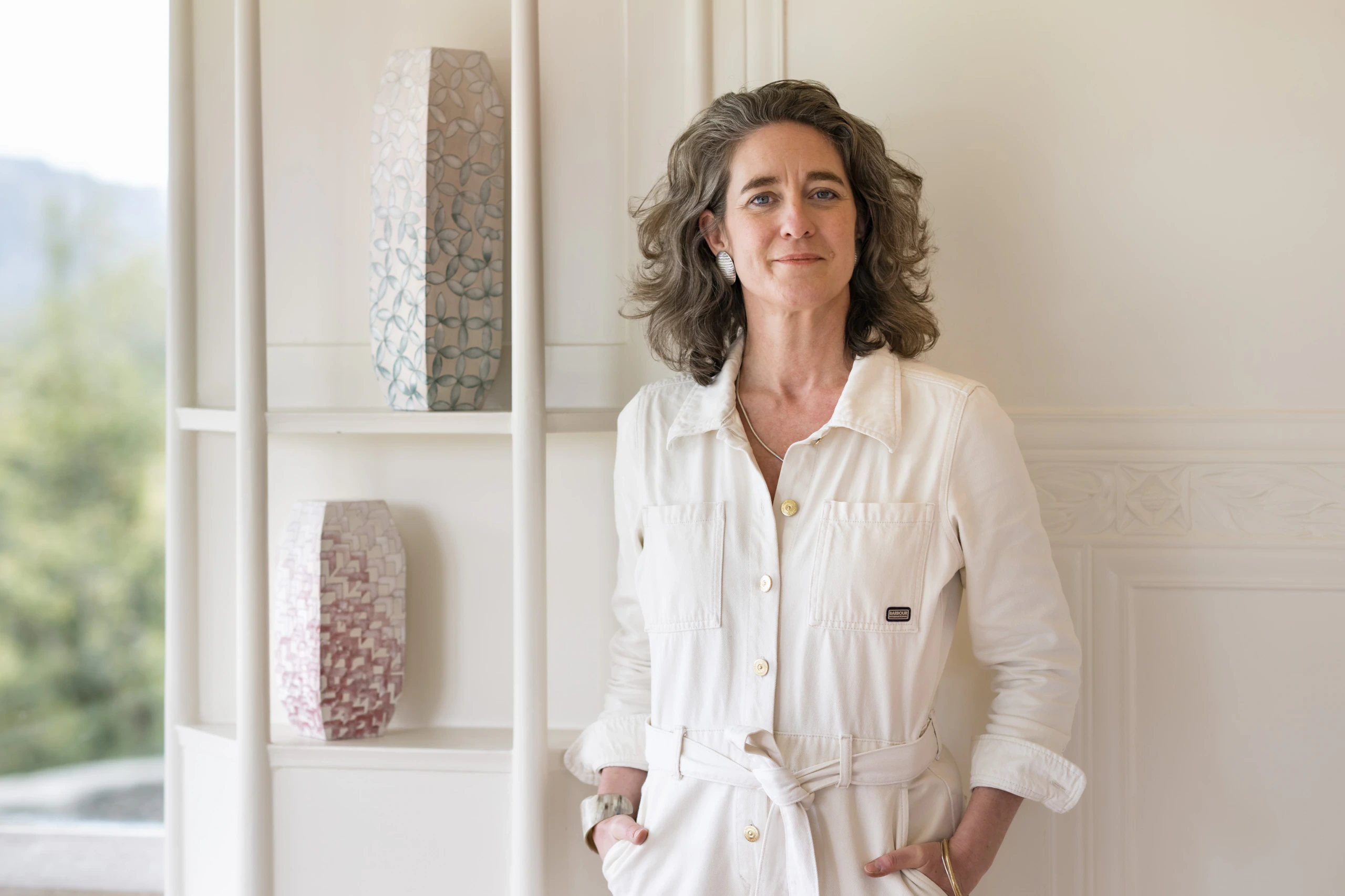 A woman with wavy brown hair wearing a white belted jumpsuit stands indoors, hands in her pockets, in front of white shelves displaying two textured ceramic vases. Natural light brightens the room.
