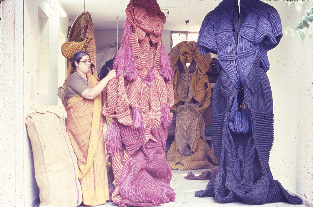 A woman in a sari stands beside large, abstract sculptures made of thick, woven fibers in shades of pink, purple, and blue, displayed in a bright indoor space.