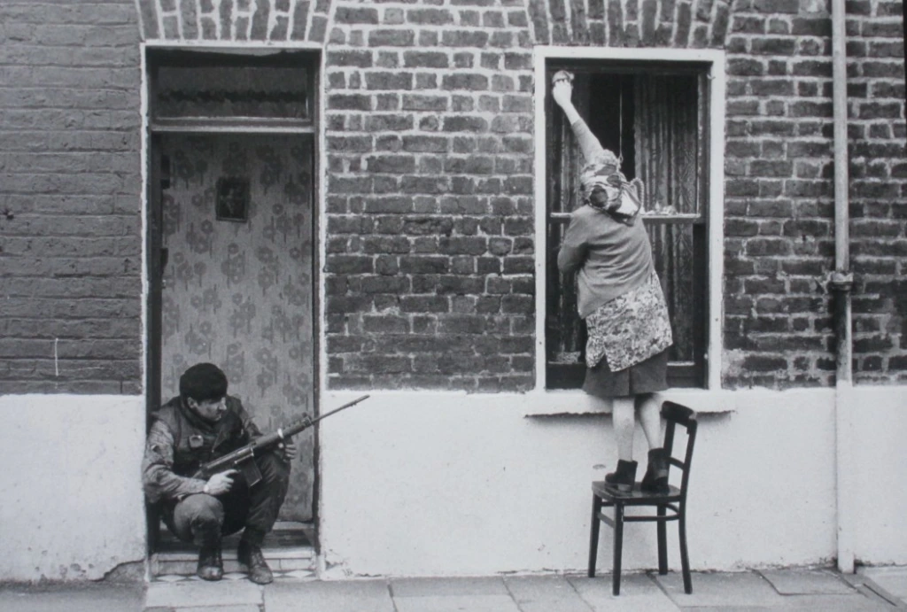 A woman stands on a chair, cleaning a window on a brick building, while a man crouches nearby in a doorway holding a rifle, partially hidden and alert.