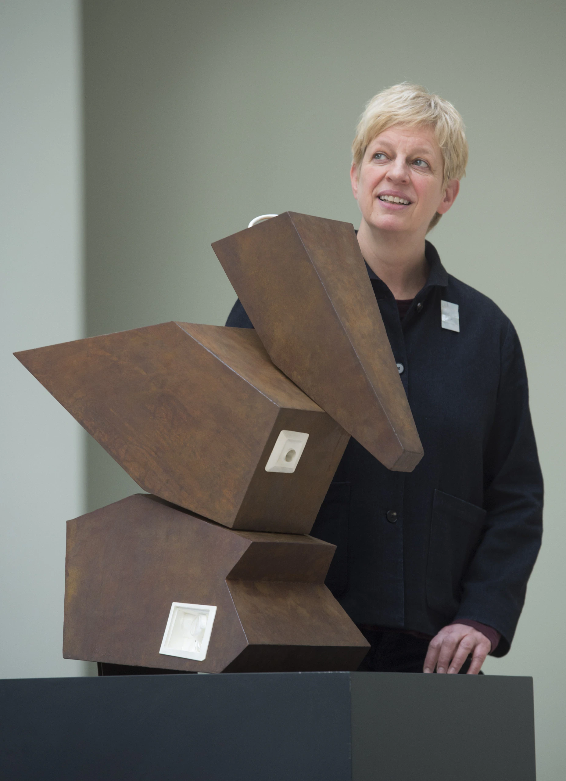 A smiling person with short blond hair stands beside a modern, abstract brown sculpture made of angular geometric shapes, displayed on a pedestal in a well-lit indoor space.