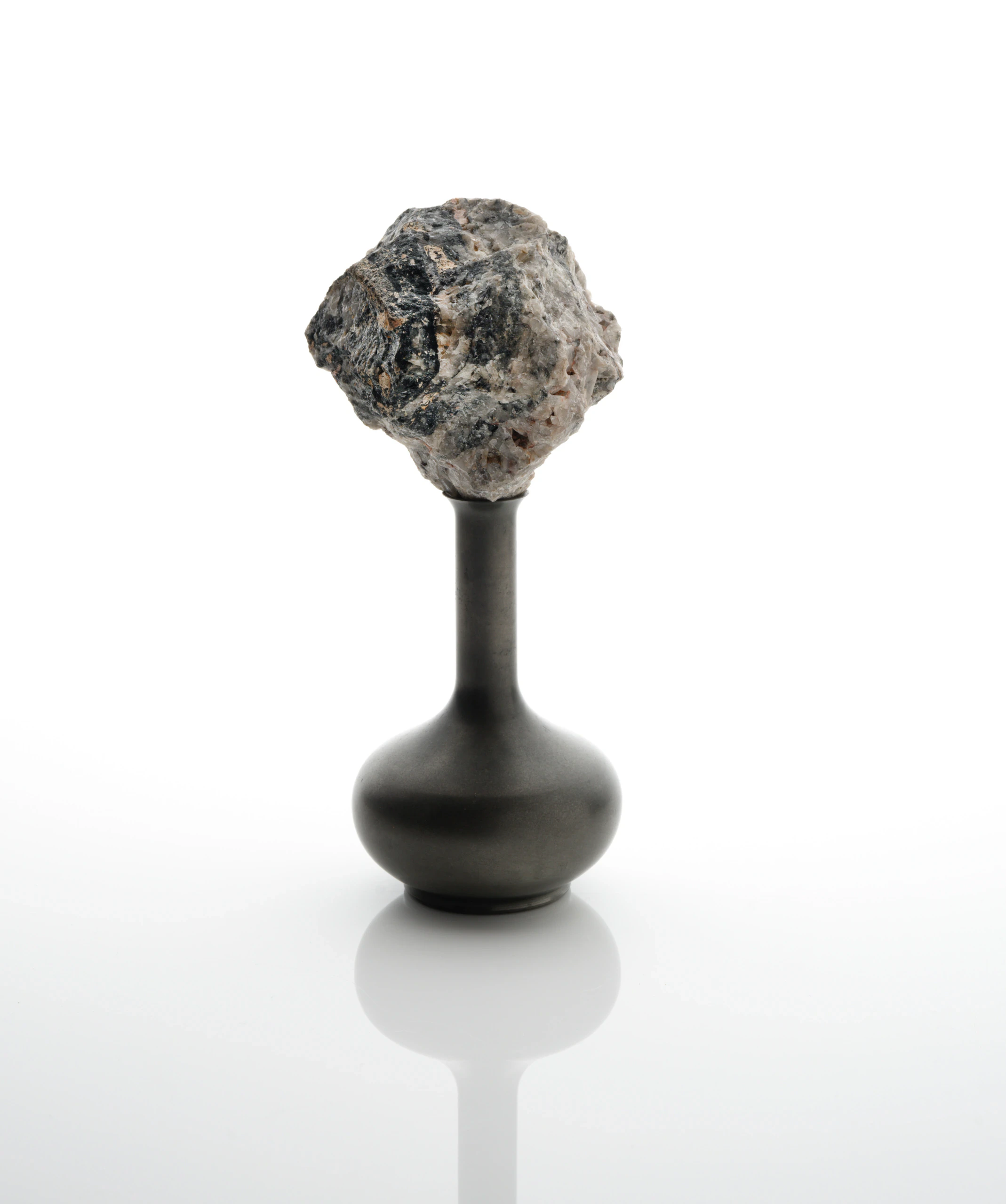 A rough, irregularly shaped rock is balanced on the narrow neck of a smooth, dark, round-bottomed vase, all set against a plain white background.