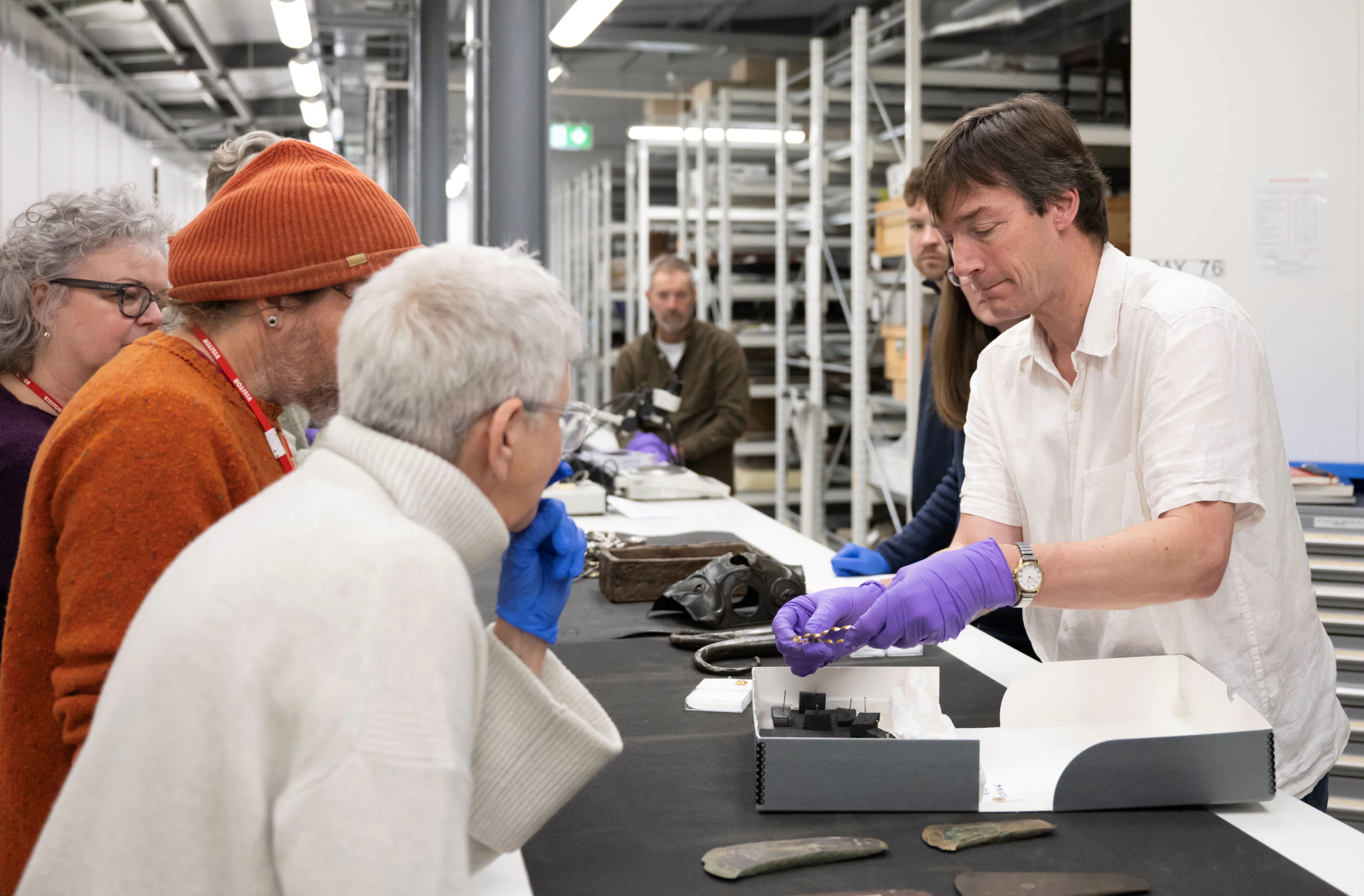 A man wearing purple gloves shows a small artifact to a group of people in a storage or laboratory setting, surrounded by shelves and various archaeological objects.