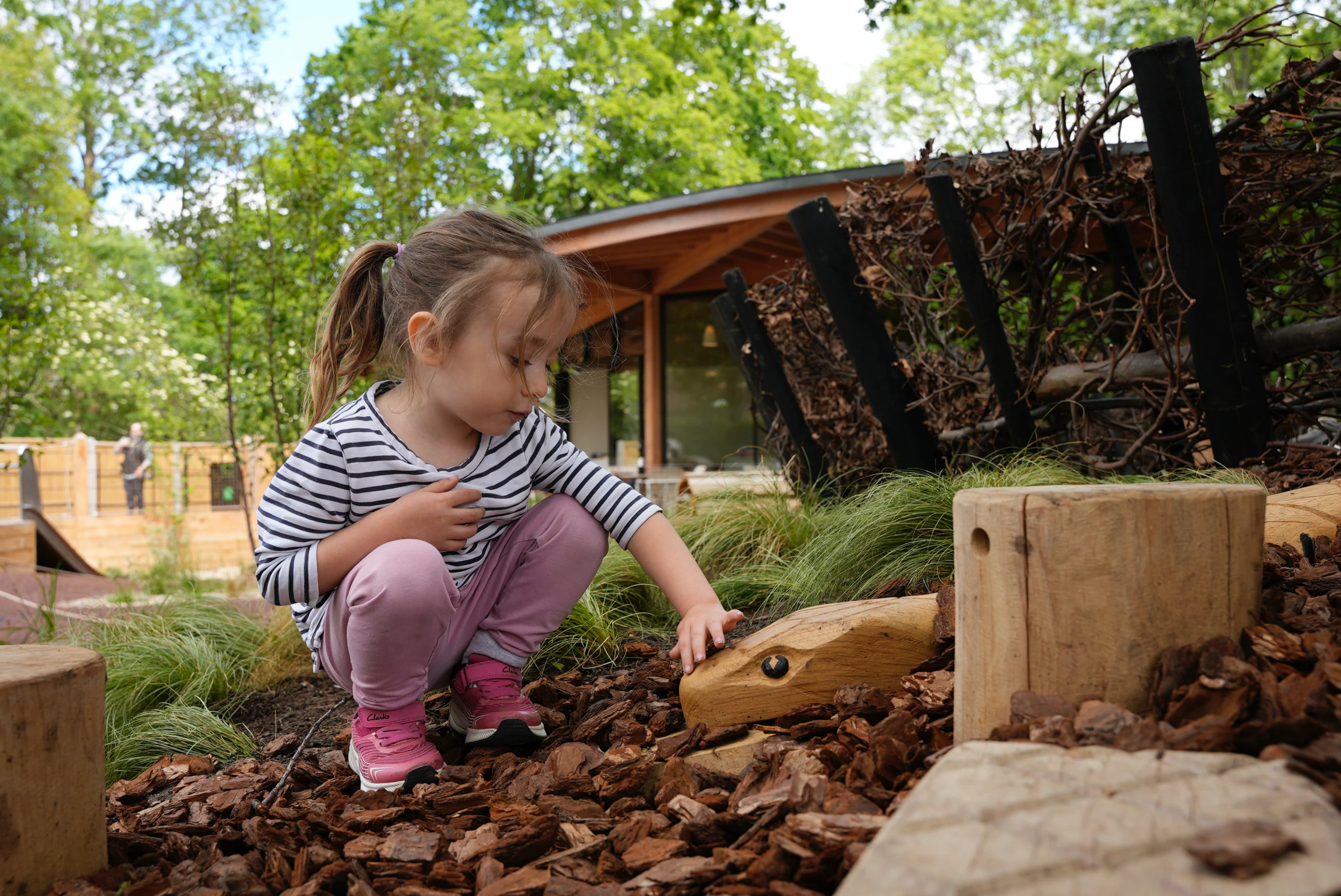 A young girl in pink pants and a striped shirt crouches outdoors, touching a wooden object on the ground surrounded by wood chips, with greenery and a wooden building in the background.