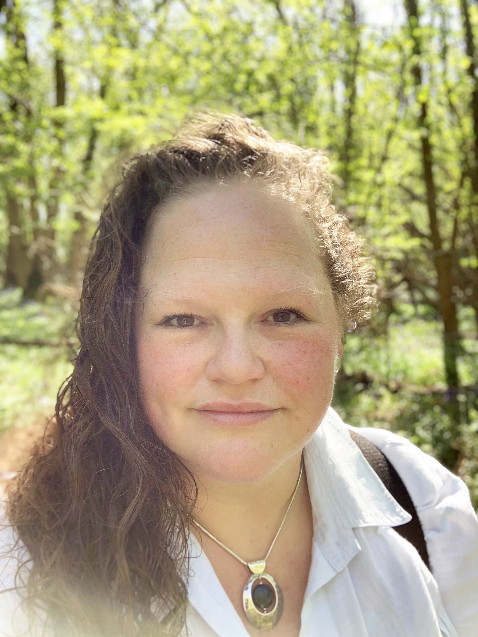 A woman with wavy brown hair and fair skin smiles slightly, wearing a white shirt and a pendant necklace, standing in a sunlit, green forest background.