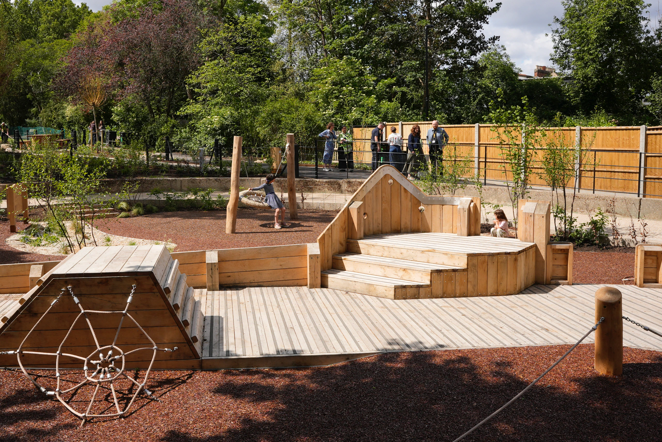 A wooden playground structure with steps, platforms, and a rope climbing area sits in a park surrounded by trees and fencing. Several people are walking or standing in the background.
