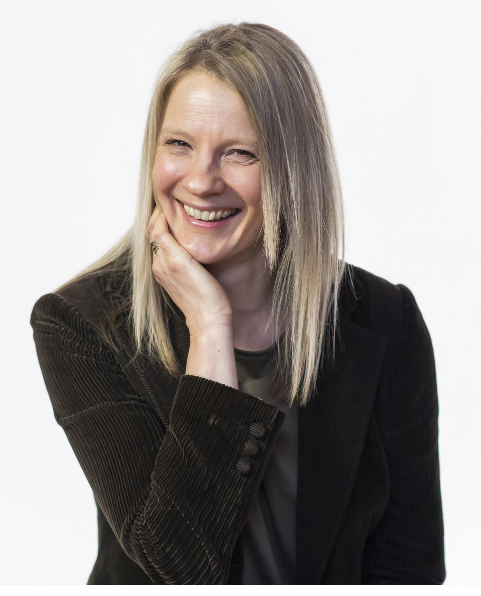 A woman with straight blonde hair, wearing a dark blazer, smiles warmly with her hand resting on her cheek against a plain white background.