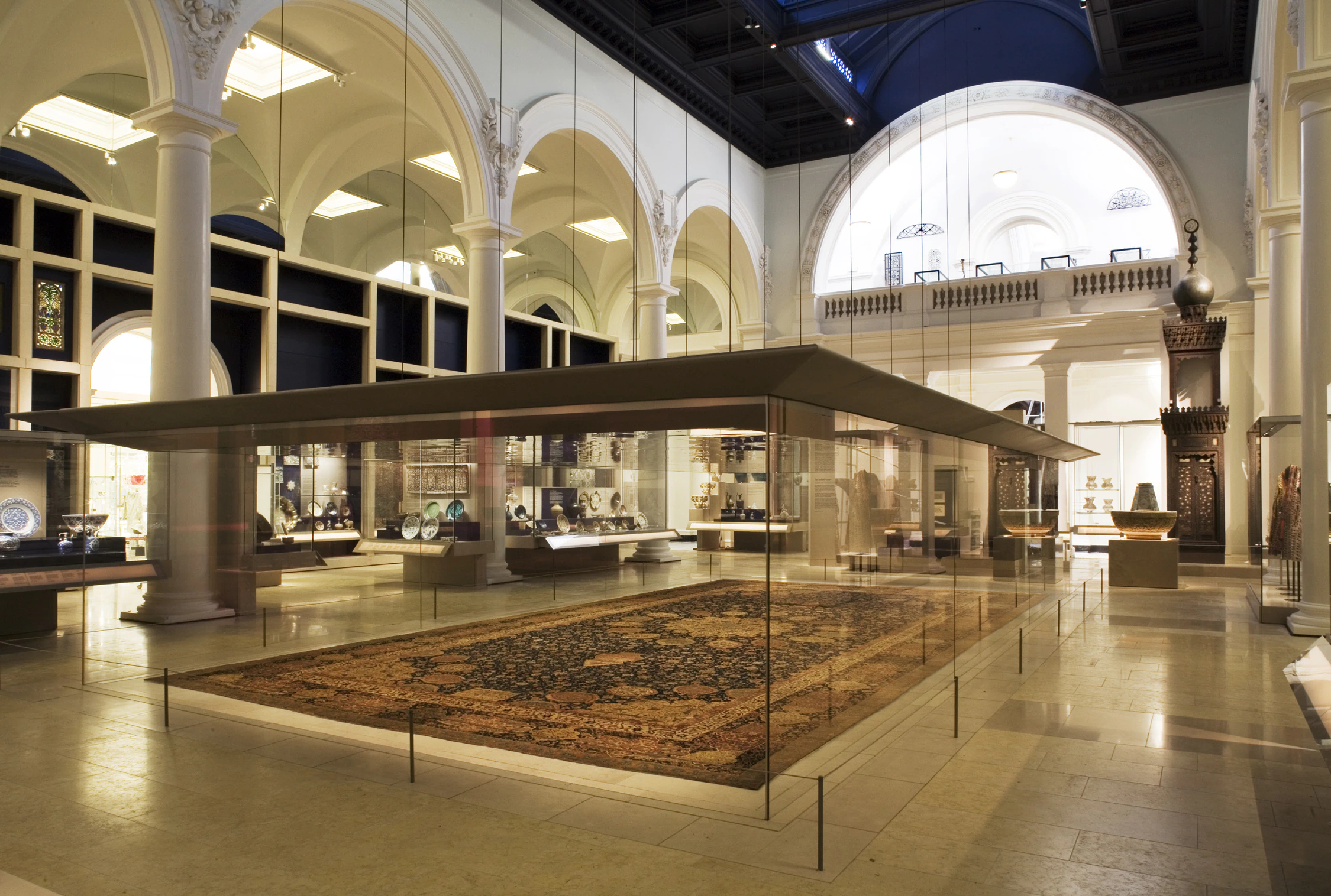 A large ornate carpet displayed in a glass case at the center of a grand museum gallery with high arched ceilings, columns, and various artifacts along the walls.